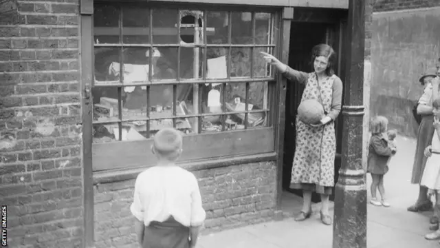 A woman holding a football and pointing out where it had smashed through the window of her shop