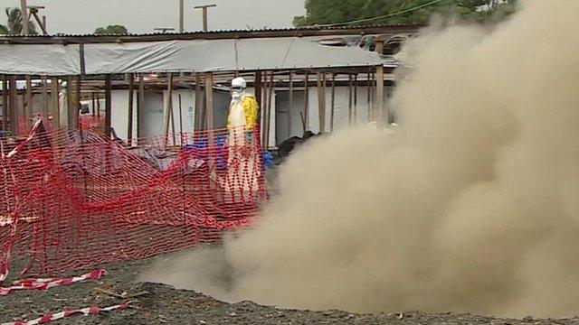 Person in protective gear watches smoke from a fire as an MSF Ebola centre in Monrovia is decommissioned