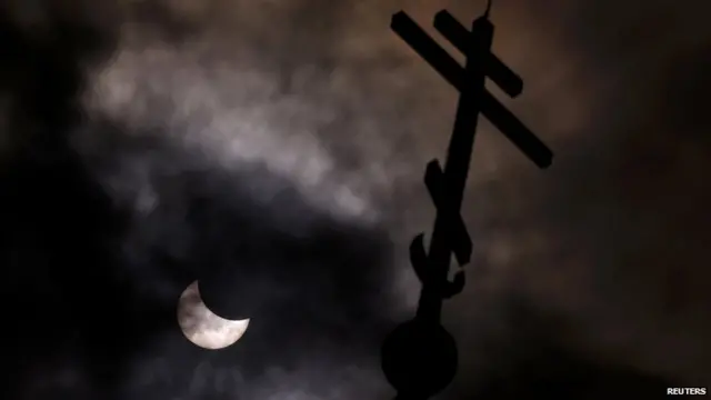 A partial solar eclipse forms in the sky through clouds near the cross of the Church of St Nicholas the Miracle-Maker in Sofia