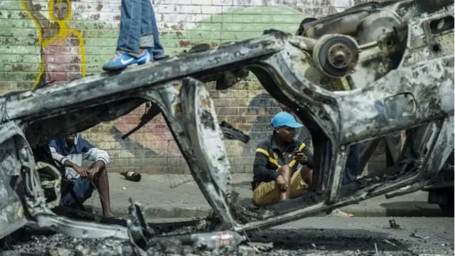 People sit next to a burnt car in the Jeppestown area of Johannesburg