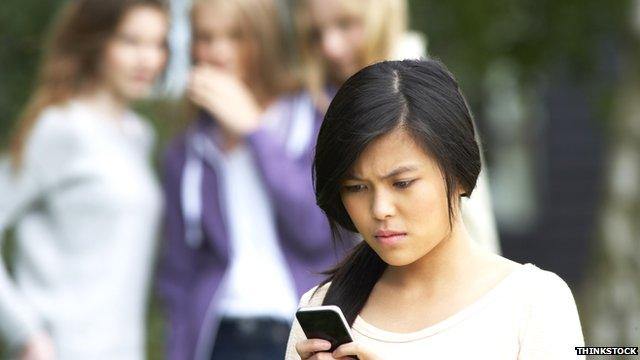 Girl gets bullying text on phone while three girls behind her look on