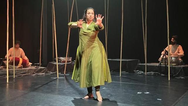 Urja Desai Thakore rehearsing for a dance show, she is wearing a green outfit, with bare feet, and she is holding both her arms up. Two men are in the distance playing musical instruments. She is dancing in a room with a black floor and black walls. 