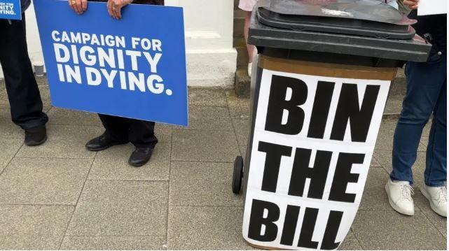 Signs for and against the Assisted Dying Bill held by protesters outside the Tynwald buildings. One sign, in favour of the bill, has the words Campaign for Dignity in Dying on a blue background. Another sign, taped onto a brown wheelie bin, has the words Bin the Bill in large black lettering.