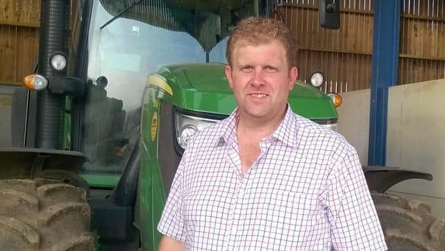 Mr Maughan, dressed in a short-sleeved checked-shirt, is standing in front of a green tractor parked in a open-sided barn