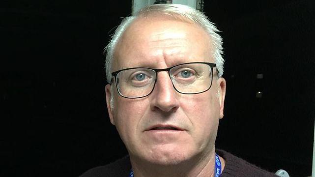 Close up of Glenn, who has white hair and black-rimmed glasses. He is standing or sitting inside with a black background behind him.