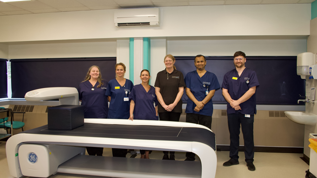 Four women and two men in medical scrubs stand in a line and smile at the camera. They are in a medical room, with a scanner in front of them.