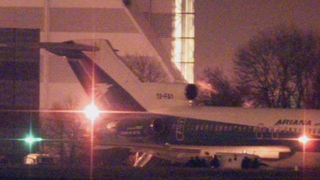 A grainy image, taken at night, shows the rear half of an airliner on the ground. Several bright lights can be seen, and several figures in silhouette are standing on the ground beneath the aircraft. There are more figures behind the aircraft, standing behind what appear to be two Land Rovers.