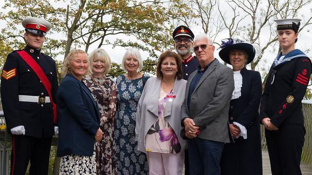Nine people are stood in a row, all smiling at the camera. The people at the ends of the row, including two others, are in ceremonial outfits, while the others are dressed in suits and dresses. They're on a bridge surrounded by woodland.
