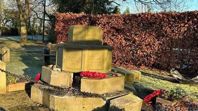 A war memorial, with a hexagonal stone base, and four tiers up to the base for the cross, though the cross is lying on the grass behind it. Some large stones are dislodged. There are Remembrance wreathes on top of, and next to, the monument.