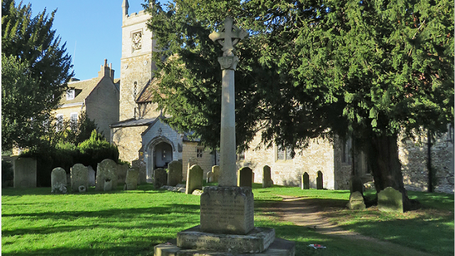 Grey stone war memorial in churchyard, surrounded by lawn, gravestones, a large tree and church in background 