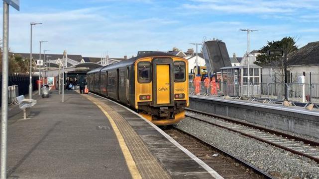 The view a platform at Newquay train station. On the left-hand side is a green and yellow Great Western Railway train. The other side is another platforms with barriers along it, and people in orange hi-vis. There is a sign on the left-hand sign with the word 'Newquay'.
