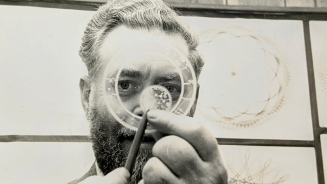 A black and white photograph of Denys Fisher in a suit holds a Spirograph drawing tool and pen close to the camera, appearing to demonstrate its use. Behind them, several intricate Spirograph designs are displayed on a wall.