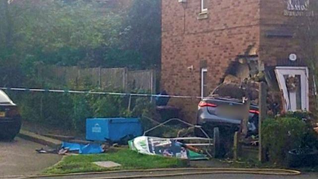 The side of a house with the rear end of a metallic blue car sticking out from a hole in the ground floor with the front door just beside it