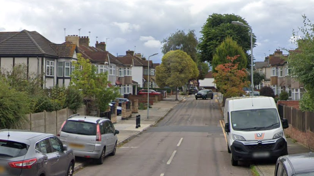 Residential street with parked cars, semi-detached houses, trees, and a white learner van.