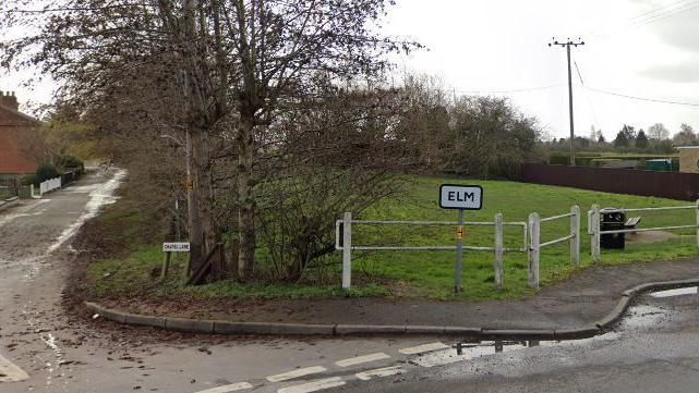 A single track residential road on the left and a sign saying Elm in front of white railings and behind it a grassy field