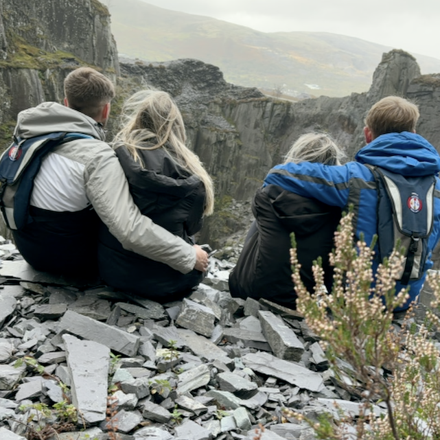 Two couples sitting high up in a slate quarry. All have their backs to the camera - with the couple on the right - a young man with short brown hair wearing a hooded grey and white coat and small blue rucksack. His arm is around the waist of a woman, with long blonde hair and a dark down style coat. Next to her on her right, is another woman, with silver hair, wearing a dark coat. A man with a blue and black jacket and small rucksack has his arm draped across the back of her neck. In front of them is a drop - and on the other side of the drop rises a wall of slate rock, with high hills in the distance