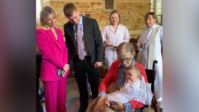 Sarah is in a wheelchair holding her grandchild surrounded by members of her family in a church