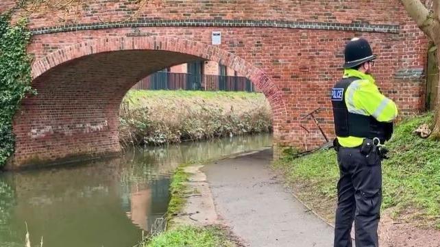 A uniformed police officer stands next a canal with a brick bridge