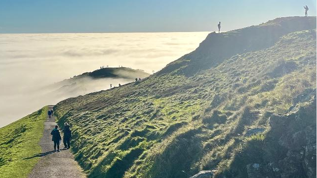 People are walking on a footpath amid the hills as well as on the top of them. Cloud surrounds the hills below but blue skies can be seen.