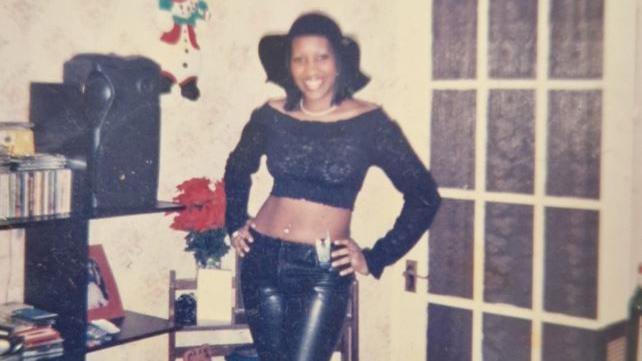 A woman in a black crop top, black floppy hat and black leather trousers smiles at the camera with her hands on her hips. Sepia toned photograph.