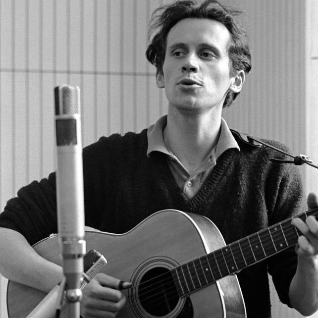 Black and white photo of Archie Fisher taken in 1963. He is standing at a microphone and is playing an acoustic guitar. He has short, wavy hair and is wearing a shirt under a dark-coloured V-neck jumper.
