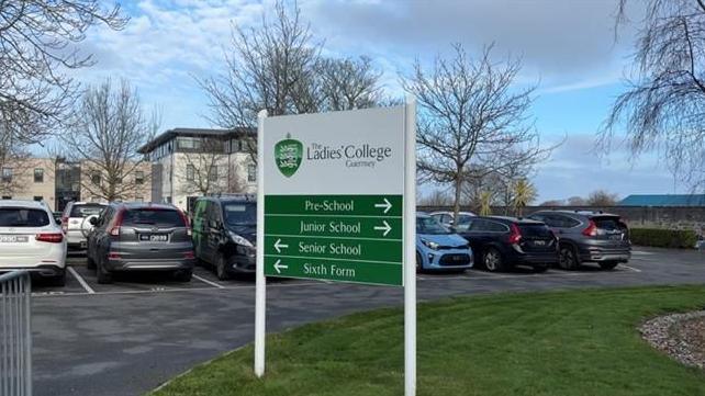 A white and green sign held up with two pillars reads THE LADIES' COLLEGE GUERNSEY. Behind it is a car park which looks full and behind that is the college which appears to be three to four storeys and modern in design. 