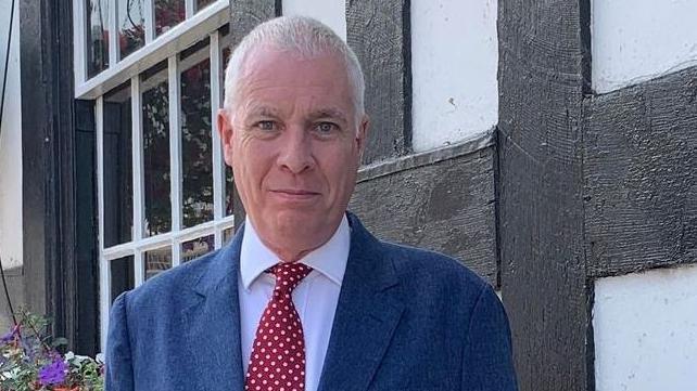 Councillor Ewen Sinclair stands outside a building with a flower bed behind him. He has grey hair and is wearing a blue suit with a jacket and tie.