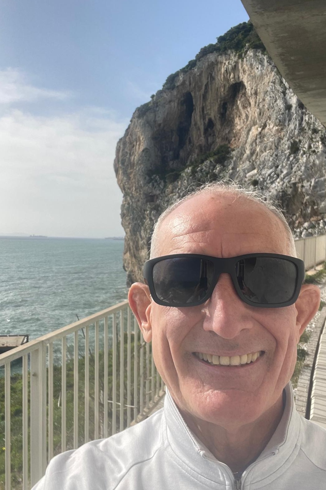Paul smiles at the camera with shades on. He has a large cliff face and sea scape behind him.