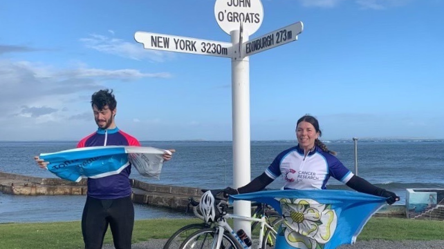 A picture of Jes Oakley holding a Yorkshire flag standing alongside her husband at the sign for John O'Groats with the sea behind.