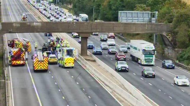 The M1 motorway, showing eight lanes of traffic, with vehicles on the road, and cars stopped. There are fire engines, ambulances and police vehicles on several lanes.