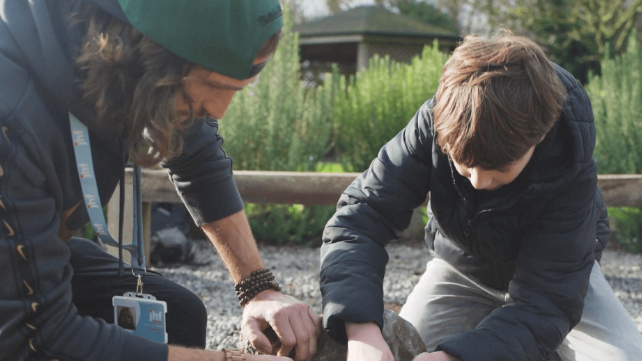 Craig, wearing a baseball cap the wrong way round and a lanyard, is kneeling outdoors on a gravelled area beside a child aged about 10, and both of them are working on something out of shot. They are in sunshine, but autumnal clothes.
