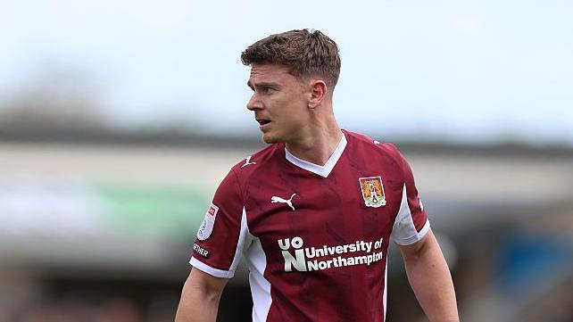 Sam Hoskins wearing a maroon Northampton Town shirt. He is looking to the side 