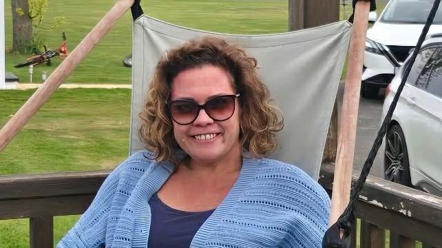 A woman wearing sunglasses and a blue jumper over a blue top sitting on a chair at a caravan park. She is smiling at the camera. 