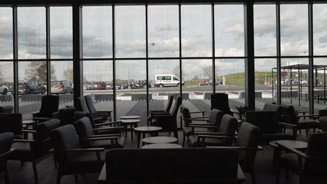 A view of what looks like a meeting lounge, with black upholstered chairs and small coffee tables, looking out through a bank of glass windows towards a car park.