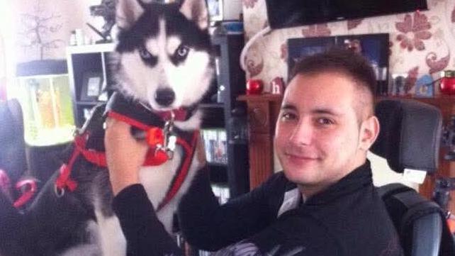 A grainy photograph of Karl Lee, in an electrically-powered wheelchair with a dog on his lap. Mr Lee and Karl are both facing the camera. Mr Lee appears to be in a living room. A fireplace can be seen in the background.