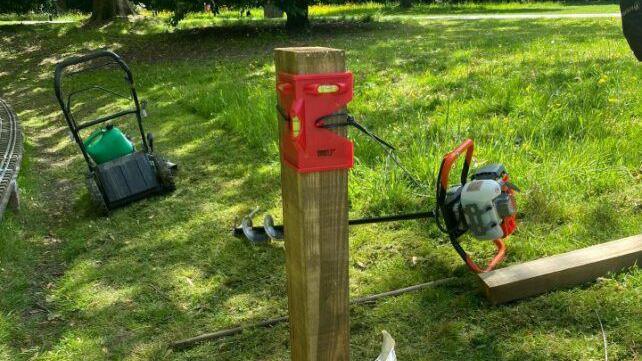 A fence post driven into the grass in Goffs Park with a lawnmower and a power ground drills next to it. A second fence post also lies on the ground waiting to be installed.