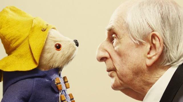 Novelist Michael Bond to the right of the image - he is wearing a suit and has white hair - and a teddy of Paddington Bear to the left in a blue coat and yellow hat. The bear and Bond are positioned so they are looking at each other - side on from the camera