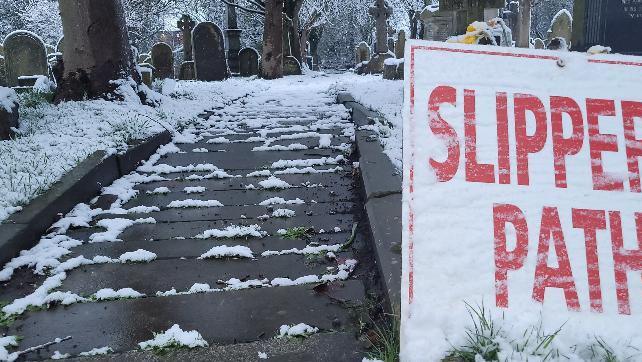 A snow-covered Slippery Path sign stands in front of a snowy graveyard