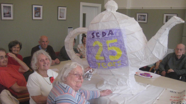  A group of older people in a large community room sitting round a table with a large teapot-shaped white lantern on it