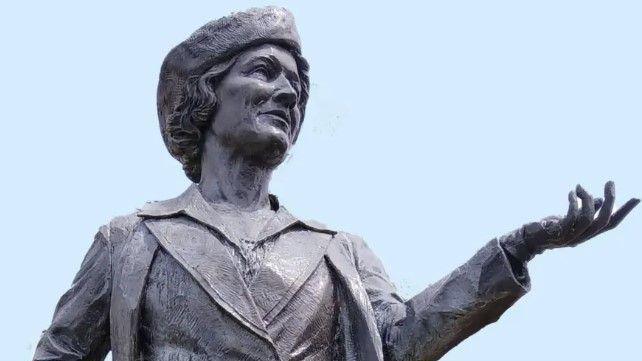 Close-up shot of a bronze statue of Nancy Astor, the first female MP to sit in the House of Commons. The statue shows Astor in a pose where her left arm is outstretched as she looks into the distance. It is a sunny day.
