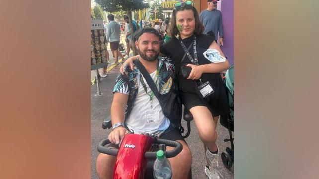 Conaire is on a red disability scooter wearing a white t-shirt and Hawaiian shirt over it and his daughter Autumn, wearing a black dress is standing beside him.
