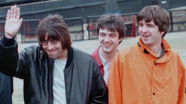 Oasis at Knebworth before their 1996 show. Liam Gallagher is waving and wearing a black leather jacket and John Lennon-style glasses, while Noel is wearing an orange jacket and also smiling. Behind them is bass player Paul McGuigan