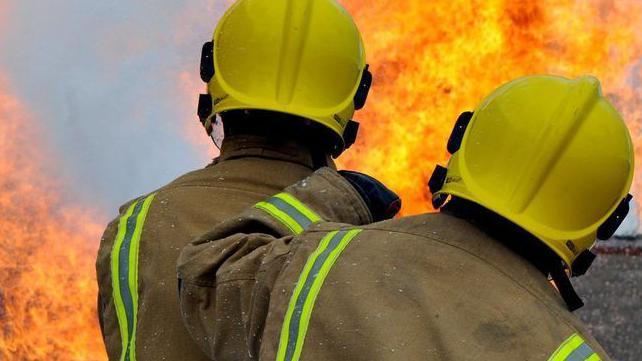 Two firefighters, wearing yellow helmets and heavy duty jackets, can be seen at the scene of a large fire.