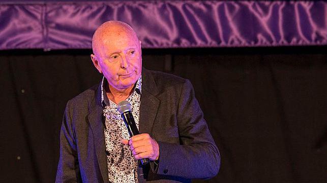 Brummy comedian Jasper Carrott performing on stage at the Henley Festival - he's wearing a dark jacket and flowery shirt and holding a microphone.