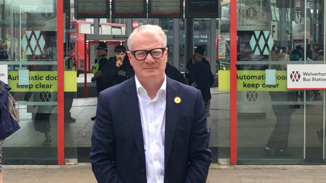 A man with white hair and black framed glasses in a blue suit and white shirt standing in front of red framed automatic signing doors with the words Wolverhampton bus station on them