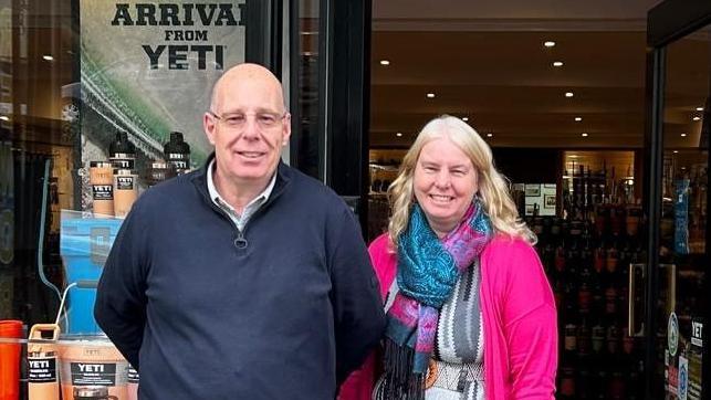 Andrew Fuller, wearing a dark blue jumper with zipped top and shirt underneath, next to his sister Liz, wearing a bright pink jacket and pink and blue patterned scarf. Both are smiling as they stand outside the front of their shop, SC Fuller, with outdoor merchandise visible