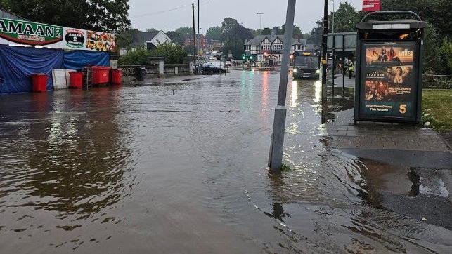 A flooded road with a bus stop and wheelie bins visible, there is a bus as well as various buildings in the background