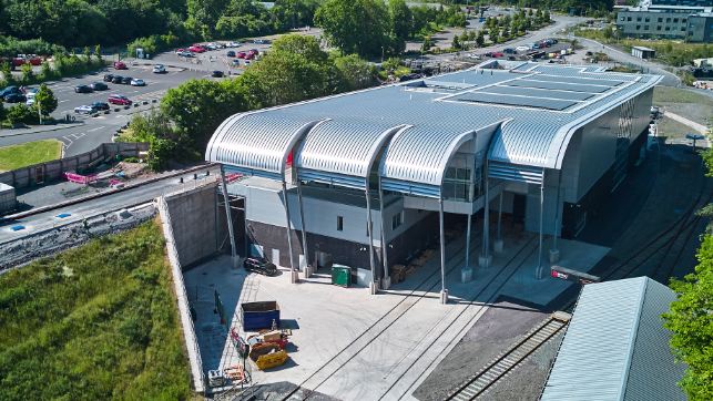 An aerial view of a tall, modern grey roofed building. There is a small railway line running out from the bottom right of the picture. In the distance are car parks, trees and other buidings