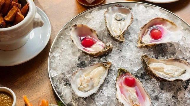A wooden table laid out with food and drink. At the centre is a round metal tray filled with crushed ice, holding six oysters in their shells. Surrounding the tray are various other food dishes.