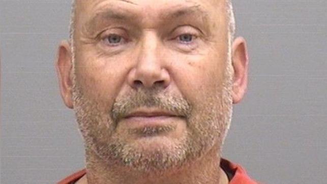 A police custody photo of a bald man with grey stubble and wearing an orange sweatshirt. The photo is taken against a plain grey backdrop and he is looking, unsmiling, at the camera.